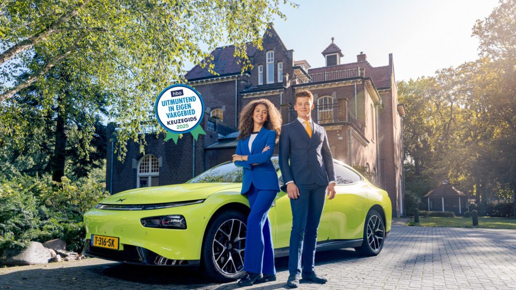 Twee IVA-studenten in blauwe pakken poseren zelfverzekerd voor een felgele elektrische auto, voor gebouw Sterrebosch op het IVA-terrein. Een blauwe zegel op de foto vermeldt 'hbo Uitmuntend in eigen vakgebied - Keuzegids 2025.