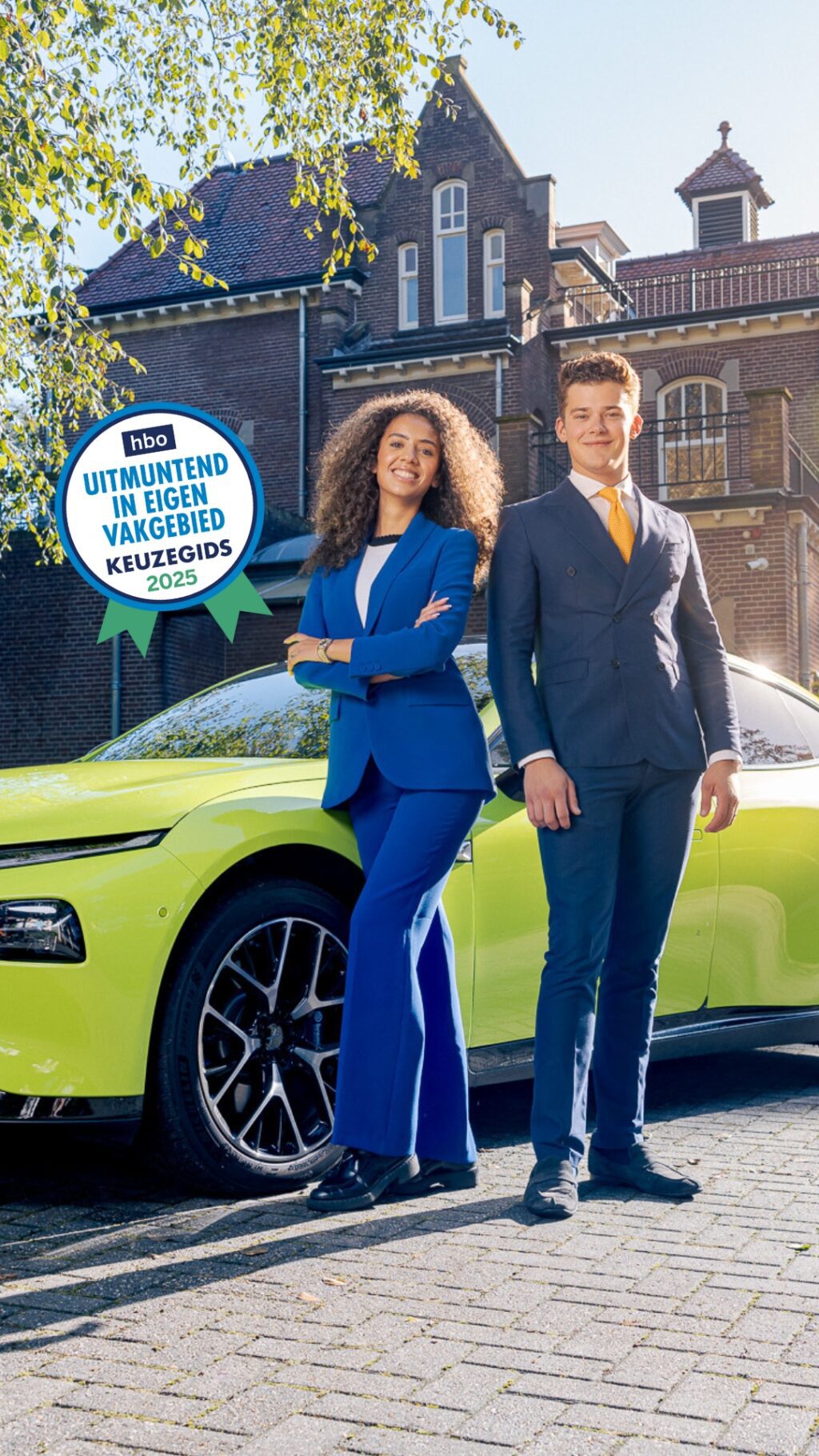 Twee IVA-studenten in blauwe pakken poseren zelfverzekerd voor een felgele elektrische auto, voor gebouw Sterrebosch op het IVA-terrein. Een blauwe zegel op de foto vermeldt 'hbo Uitmuntend in eigen vakgebied - Keuzegids 2025.
